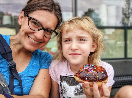A woman and a girl smile together, with the girl holding a chocolate donut topped with colorful sprinkles.