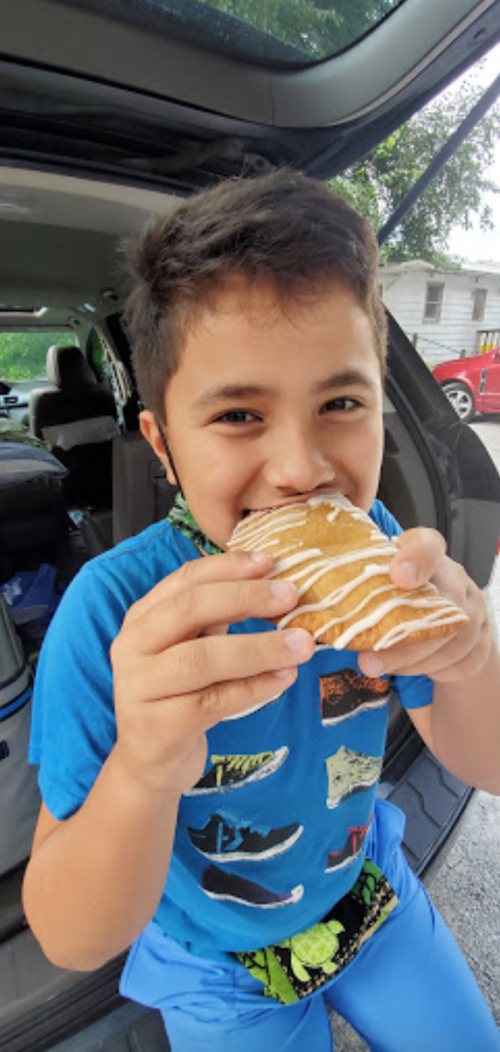 A young boy in a blue shirt smiles while holding a pastry with icing, sitting in a car.