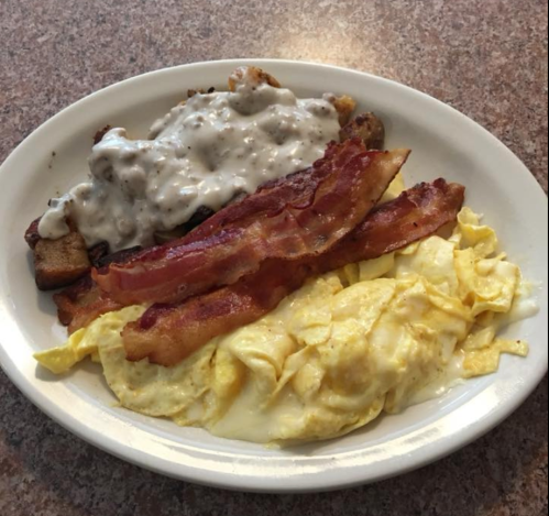 A plate of breakfast featuring scrambled eggs, bacon, and biscuits topped with sausage gravy.