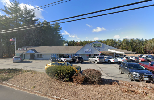 A small grocery store with a parking lot, surrounded by trees and a clear blue sky. American flag visible.