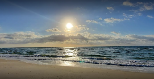 A serene beach scene at sunset, with gentle waves and a bright sun illuminating the sky and water.