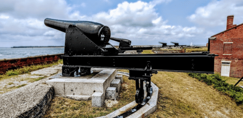 A historic cannon on a stone base, overlooking a grassy area with a cloudy sky in the background.