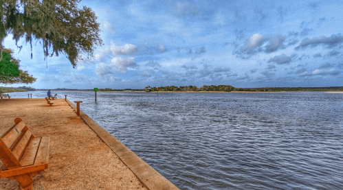 A serene riverside view with benches, trees, and a cloudy sky reflecting on the water.