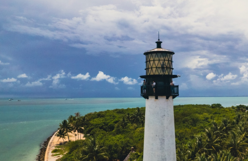 A tall lighthouse stands on a coastal landscape with palm trees, surrounded by turquoise waters and a cloudy sky.