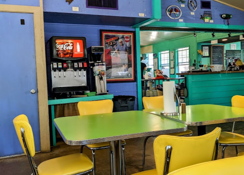 A colorful diner interior featuring a green table, yellow chairs, a Coca-Cola machine, and a vibrant blue wall.