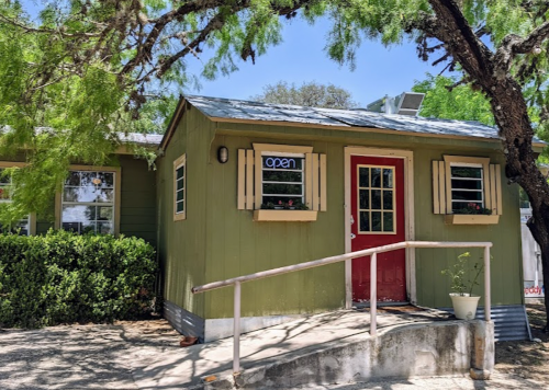A small green building with a red door and an "open" sign, surrounded by trees and plants.