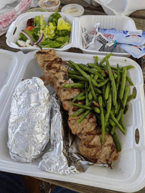 A takeout meal featuring grilled meat, green beans, a baked potato wrapped in foil, and a side salad.