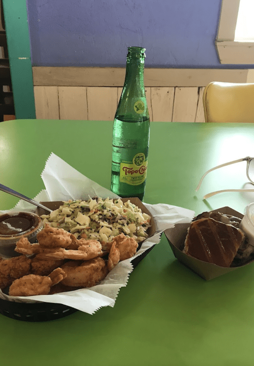 A plate of fried shrimp with coleslaw, a bottle of Topo Chico, and a dessert on a green table.