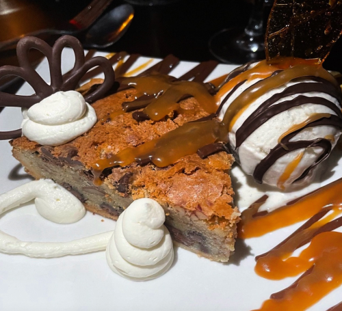 A slice of dessert with chocolate drizzle, caramel sauce, ice cream, and decorative whipped cream on a white plate.