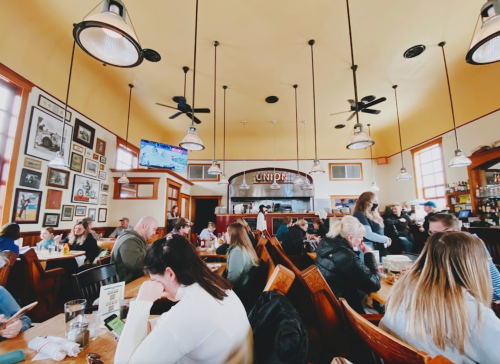 A busy restaurant interior with diners at wooden tables, vintage decor, and a visible kitchen area in the background.
