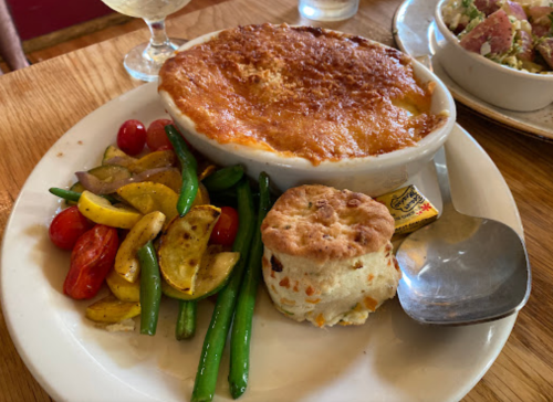 A hearty dish featuring a baked casserole, mixed vegetables, and a biscuit on a white plate.