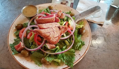 A fresh salad topped with sliced grilled chicken, red onions, tomatoes, and olives, served with a dressing on the side.