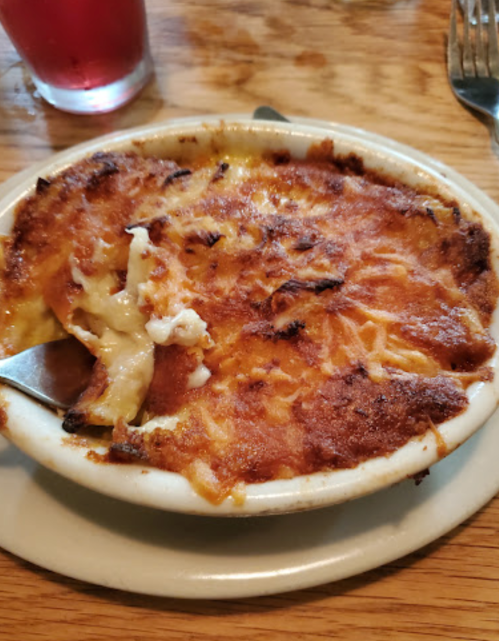 A baked dish with a golden, crispy top, served in a white bowl on a wooden table, with a spoon beside it.