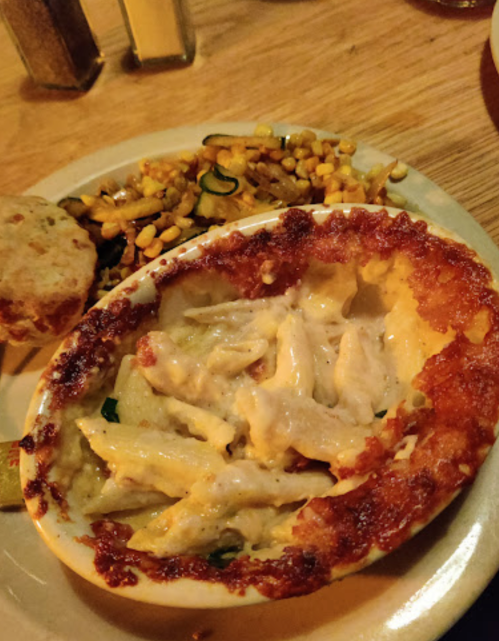 A baked pasta dish with a golden crust, served alongside a side of corn and zucchini salad and a biscuit.