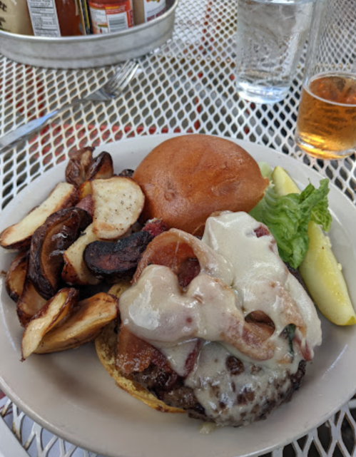 A plate featuring a cheeseburger with bacon, lettuce, and a side of roasted potatoes, served with a pickle and a drink.