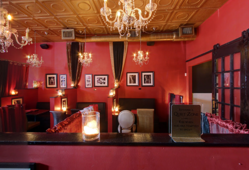 Cozy restaurant interior with red walls, chandeliers, and framed photos, featuring intimate seating and soft lighting.