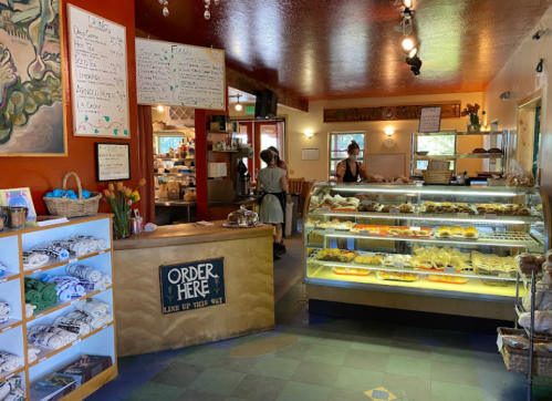 A cozy café interior with a display case of pastries, a counter labeled "Order Here," and staff preparing orders.