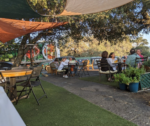 Outdoor dining area with tables under shade, surrounded by greenery and a food truck in the background.