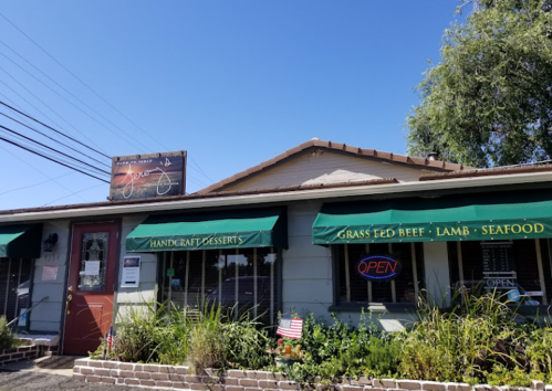 A cozy restaurant with green awnings, a sign, and an "OPEN" neon light, surrounded by plants and blue sky.