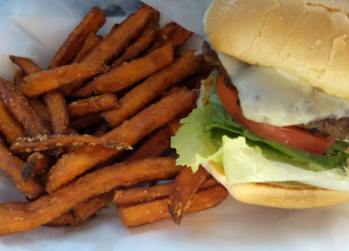 A close-up of a cheeseburger with lettuce, tomato, and a side of crispy sweet potato fries.