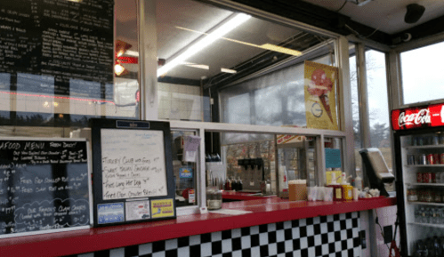 A retro-style food stand with a menu board, condiments, and a Coca-Cola fridge in a cozy setting.
