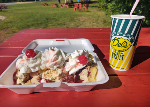 A takeout container with ice cream topped with whipped cream and cherries, next to a striped cup of drink on a picnic table.