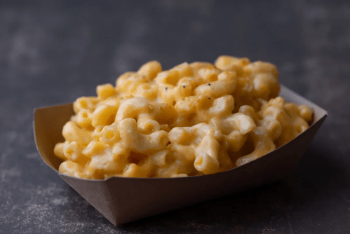 A serving of creamy macaroni and cheese in a brown paper tray on a dark surface.