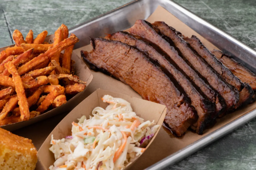 A metal tray with sliced brisket, sweet potato fries, coleslaw, and cornbread on a rustic wooden surface.