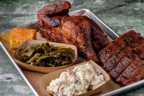 A metal tray with smoked chicken, ribs, collard greens, potato salad, and cornbread.