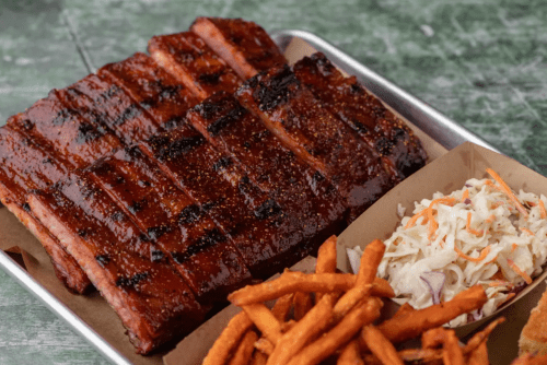 A platter featuring grilled ribs, sweet potato fries, and coleslaw on a rustic surface.