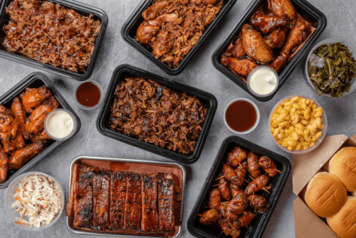 A variety of barbecue dishes including wings, pulled pork, sides, and rolls arranged in black containers on a gray surface.
