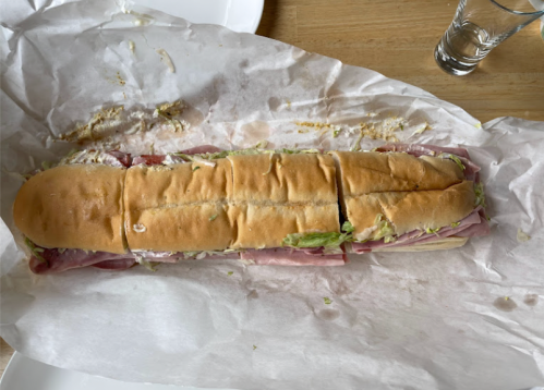 A long sub sandwich with layers of ham, lettuce, and other fillings, wrapped in white paper on a wooden table.