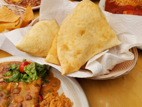 Two golden, crispy fried pastries on a paper towel, accompanied by a plate of rice and a colorful bean dish.