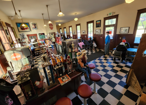 A vintage diner with checkered floors, bar stools, and patrons enjoying their meals at tables. Brightly lit and inviting.