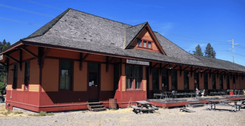 Historic train station with a red and orange exterior, surrounded by gravel and trees under a clear blue sky.