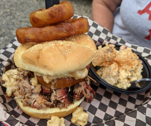 A stacked burger with pulled pork, macaroni, and onion rings, served with a side of creamy potato salad.