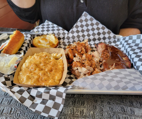 A plate with BBQ chicken, pulled pork, mac and cheese, and cornbread, served on a checkered paper.