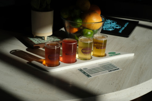 A flight of three craft beers on a white paddle, with a bowl of colorful fruits in the background.