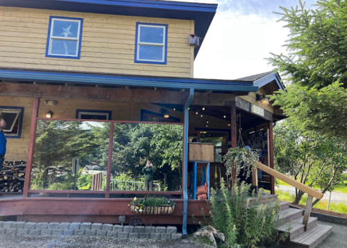A wooden building with large windows, surrounded by greenery, featuring a staircase and outdoor seating area.