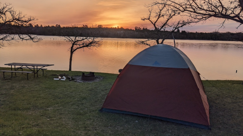 A tent by a lake at sunset, with trees and a picnic table in the background.