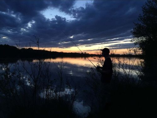 A silhouette of a person fishing by a calm lake at sunset, with colorful clouds reflecting on the water.