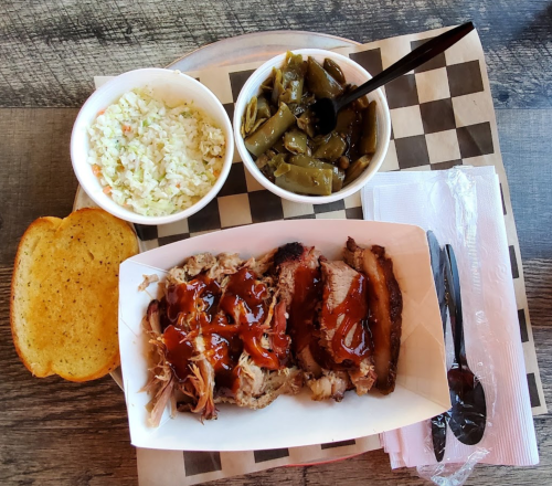 A plate of barbecue meat with sauce, served with coleslaw, green beans, and a slice of toasted bread.