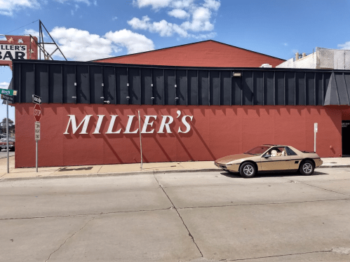 A beige car parked in front of a red building with "MILLER'S" painted on the wall. Blue sky with clouds above.