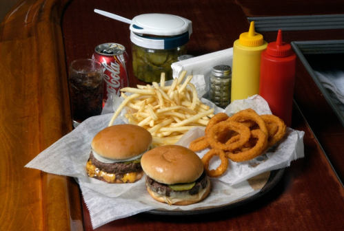 A platter with two burgers, fries, onion rings, pickles, and condiments, alongside a soda and a glass of cola.