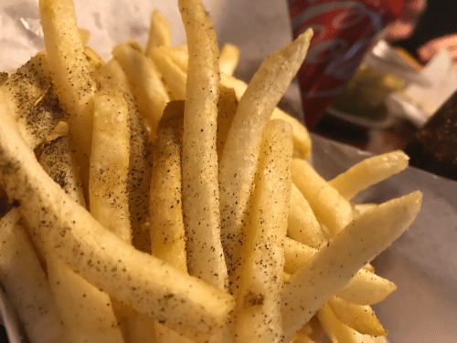 A close-up of golden French fries sprinkled with black pepper, served in a paper-lined basket.