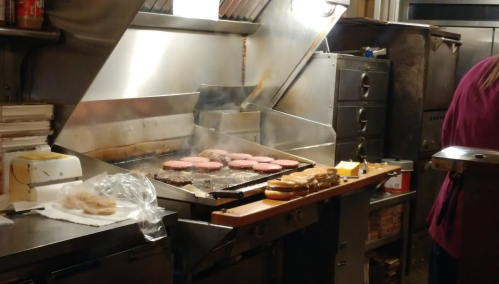 A grill with several burgers cooking, surrounded by kitchen equipment and a person in the background preparing food.