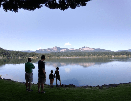 Four people stand by a lake, gazing at mountains in the distance under a clear blue sky.