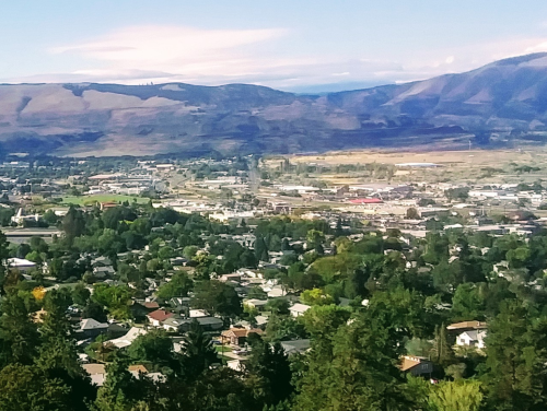 A panoramic view of a valley with houses, trees, and mountains in the background under a clear blue sky.