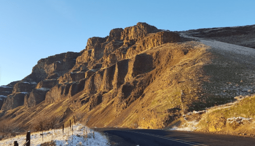 A sunlit rocky cliffside with layered formations, beside a snow-dusted road and grassy hillside.