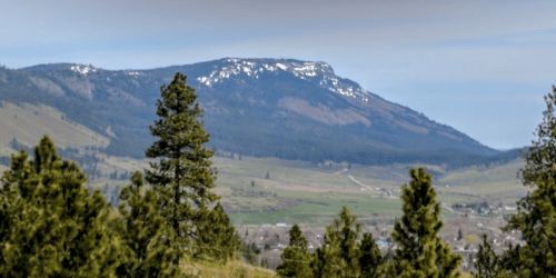 A scenic view of a mountain with snow-capped peaks, surrounded by green hills and trees under a clear sky.